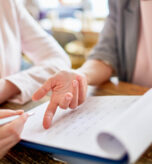 Dwie kobiety przeglądające dokumenty. Jedna z nich wskazuje palcem na kartkę, a druga trzyma długopis, gotowa do pisania. Temat: Skala semantyczna - jak ją skutecznie wykorzystać w kwestionariuszu ankiety?