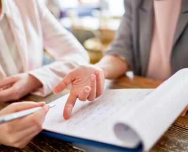 Dwie kobiety przeglądające dokumenty. Jedna z nich wskazuje palcem na kartkę, a druga trzyma długopis, gotowa do pisania. Temat: Skala semantyczna - jak ją skutecznie wykorzystać w kwestionariuszu ankiety?