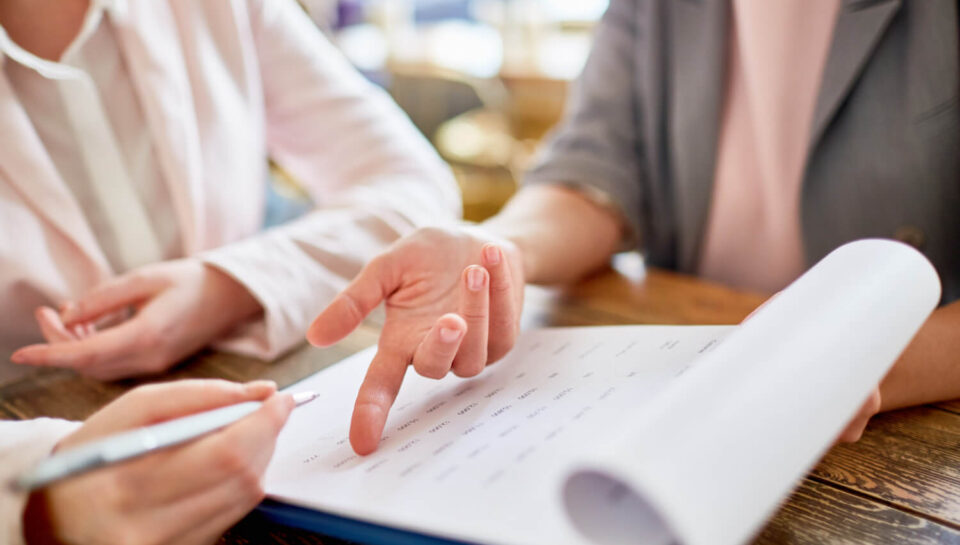 Dwie kobiety przeglądające dokumenty. Jedna z nich wskazuje palcem na kartkę, a druga trzyma długopis, gotowa do pisania. Temat: Skala semantyczna - jak ją skutecznie wykorzystać w kwestionariuszu ankiety?