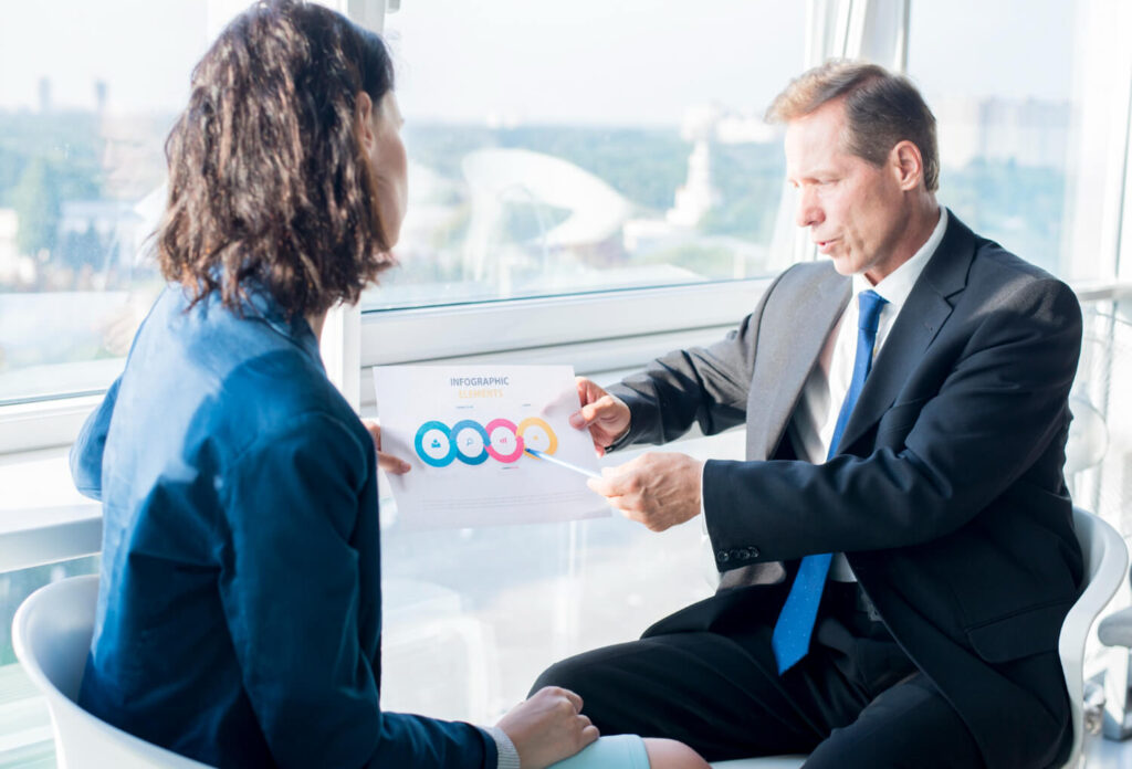 Analiza Churn Rate - co to jest i dlaczego jest kluczowy dla Twojego biznesu? 1 Mężczyzna w garniturze i kobieta w marynarce siedzą przy oknie i omawiają dokument z infografiką. Temat: Analiza Churn Rate - co to jest i jak zapobiec rezygnacji klientów?