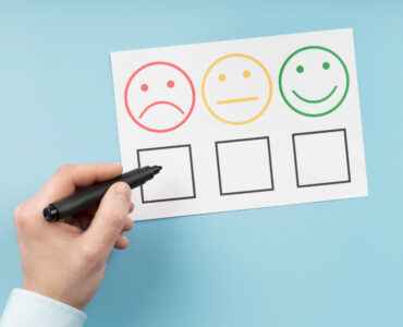 Dłoń trzymająca pisak nad ankietą satysfakcji, przygotowująca się do zaznaczenia opcji z czerwoną, smutną buźką. Temat: Wskaźnik Churn Rate