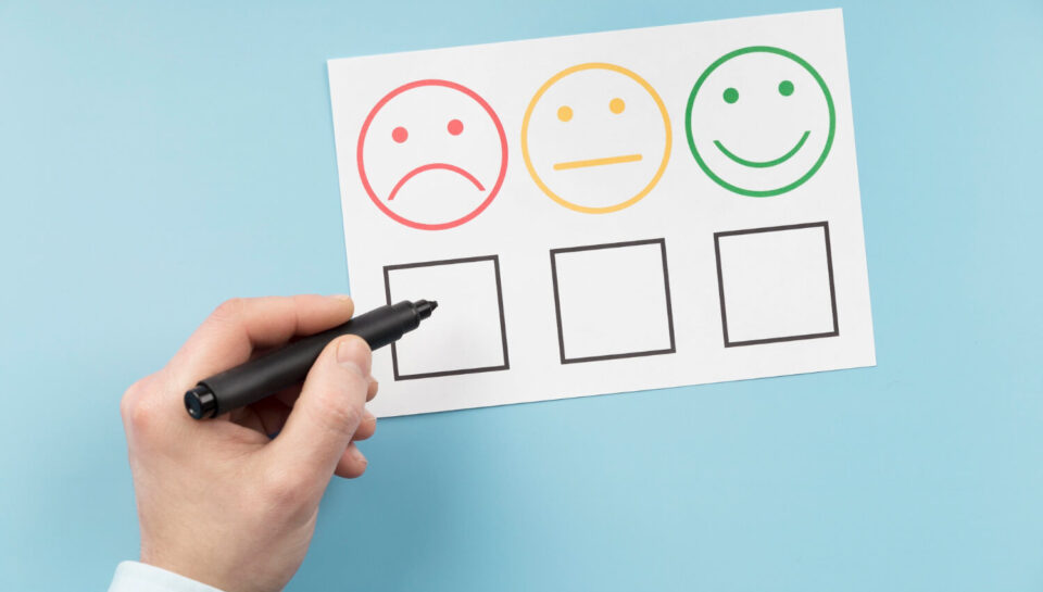 Dłoń trzymająca pisak nad ankietą satysfakcji, przygotowująca się do zaznaczenia opcji z czerwoną, smutną buźką. Temat: Wskaźnik Churn Rate