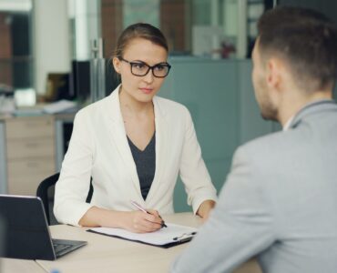 Kobieta w białym żakiecie i okularach rozmawia z mężczyzną w biurze podczas rozmowy podsumowującej wyniki