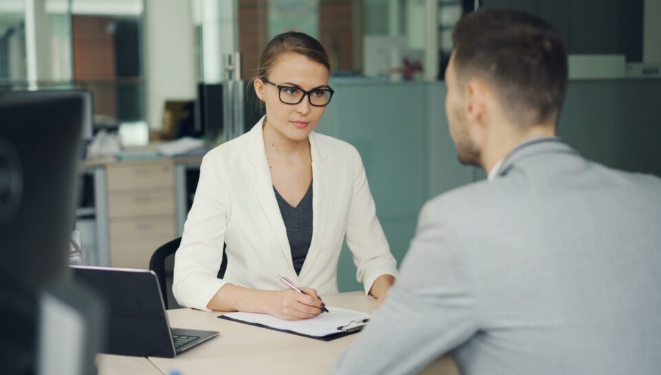 Kobieta w białym żakiecie i okularach rozmawia z mężczyzną w biurze podczas rozmowy podsumowującej wyniki